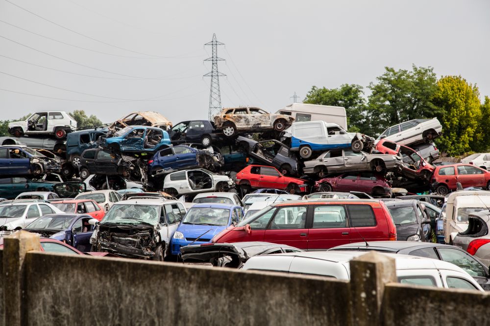 A scrapyard with numerous stacked and damaged scrap cars in Singapore, some with visible dents and missing parts, under an overcast sky with trees and a power line in the background.