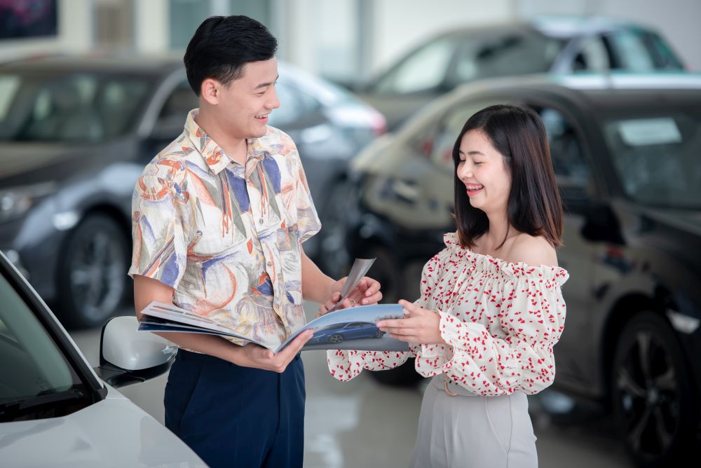 A man and woman stand by a car in a showroom, looking at a brochure together and smiling, exploring cheap car leasing Singapore options.