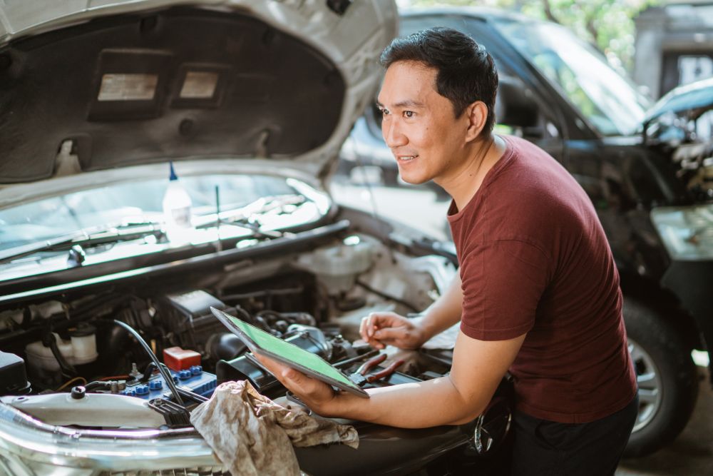 A person in a red shirt stands next to an open car hood, holding a tablet and tools, appearing to inspect or repair the vehicle—perhaps consulting some Auto Repair Tips Every Car Owner Should Know.