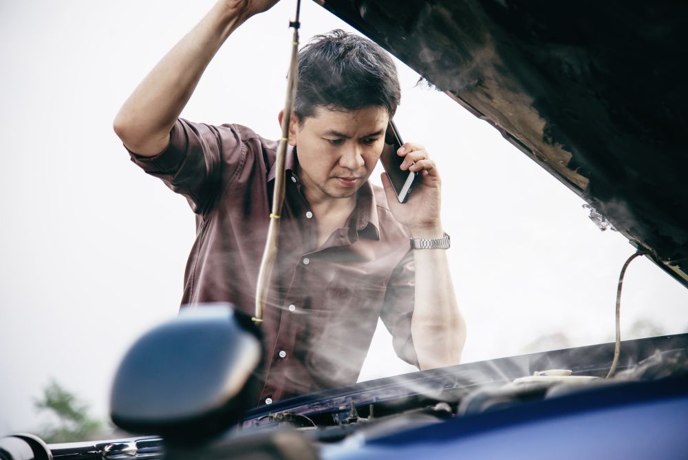 A man talks on the phone while inspecting a smoking car engine with the hood open, perhaps wondering what happens during a car gearbox overhaul as he assesses the extent of the damage.