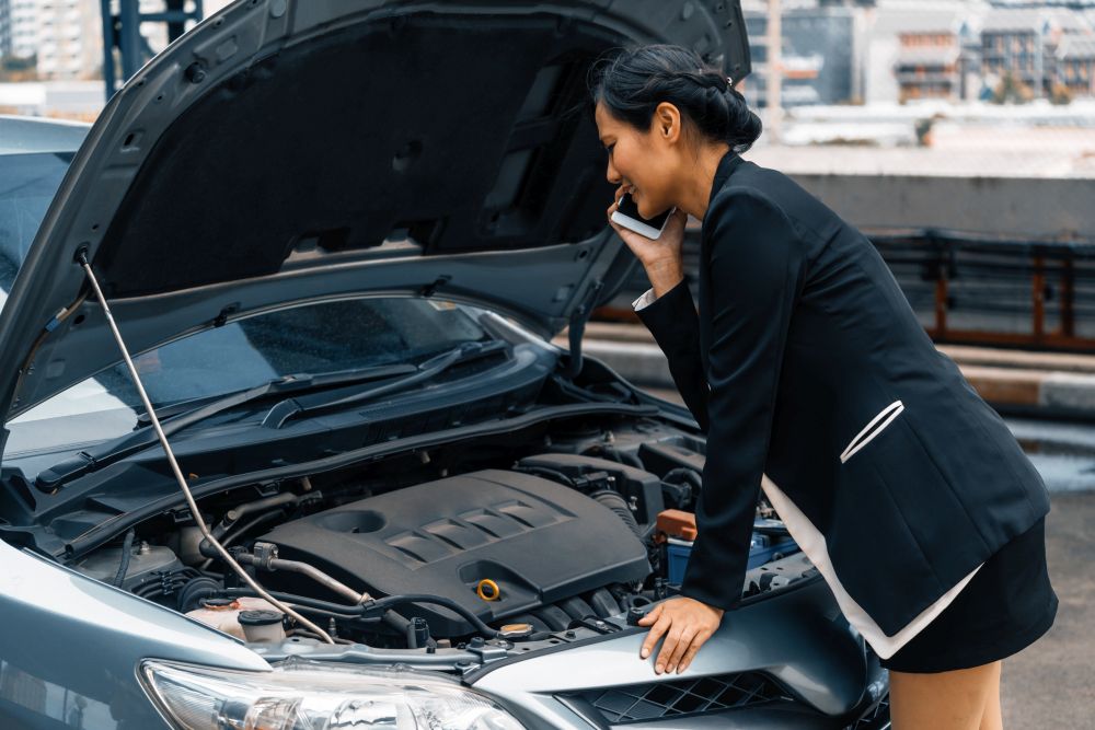 Woman in business attire stands by a car with the hood open, looking at the engine and talking on her phone, possibly seeking assistance—highlighting firsthand why regular car servicing is important.